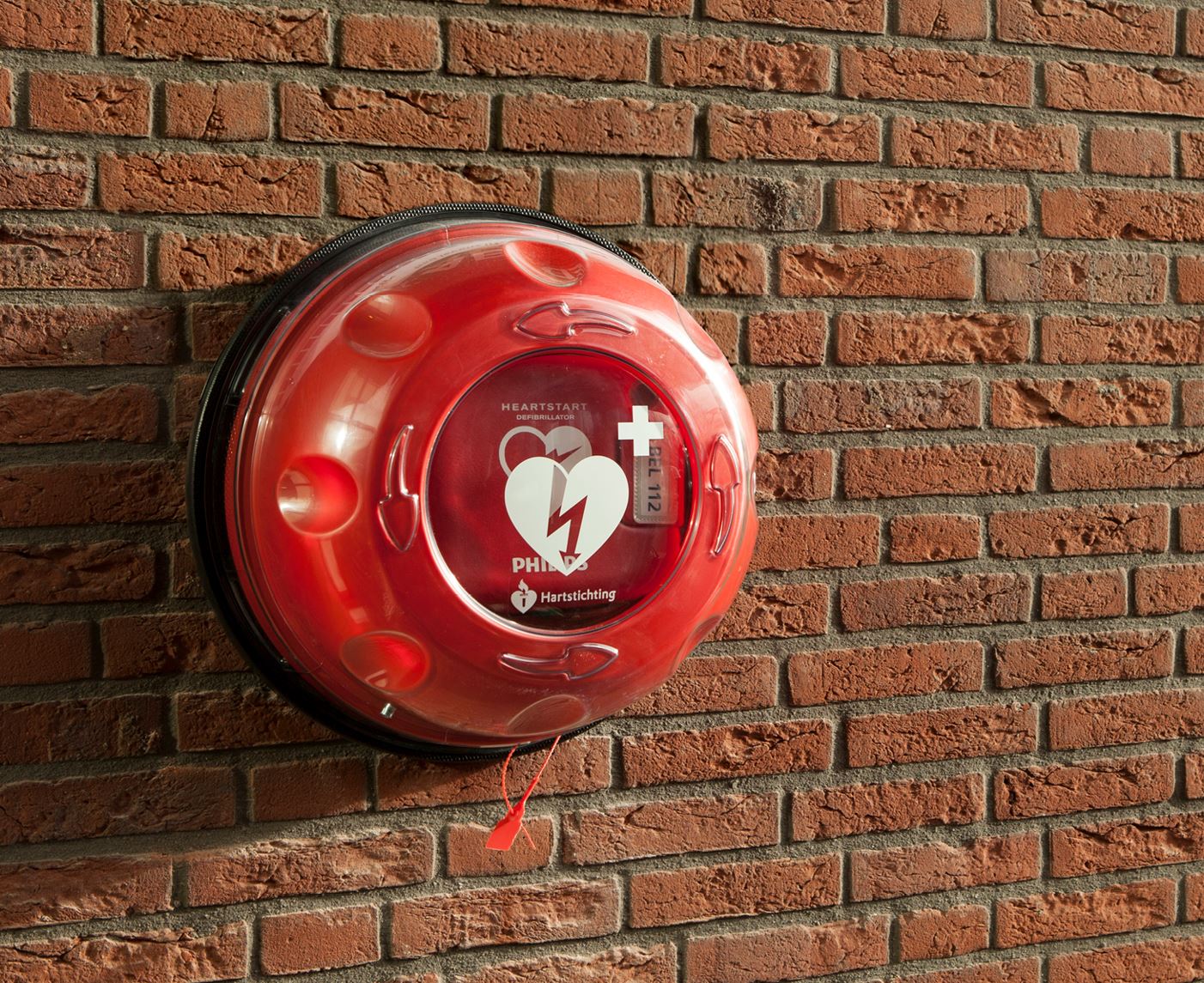 Rode AED-buitenkast aan een bakstenen muur, afbeelding uit de BuurtAED-flyer